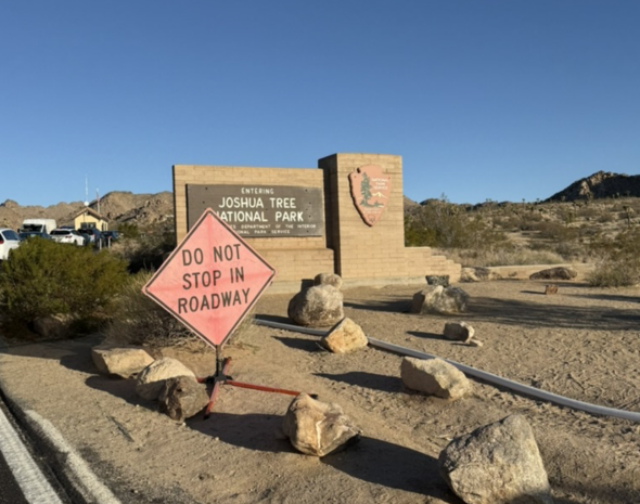 A flat desert ground with uneven rocks and bushes scattered around. In the distance stands a stone gate that says "Joshua Tree National Park."