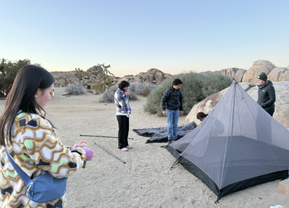 On a sandy, flat ground, a bunch of people are congregating around a tent, trying to assemble the components together.