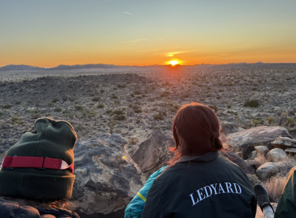 A tranquil view of a flat desert being illuminated by an intense orange sunrise in the distance. 