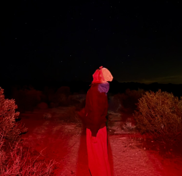 A man with his head tilted upward is admiring the dazzling sky full of stars. There are red light emitting from headlamps that's surrounding him.