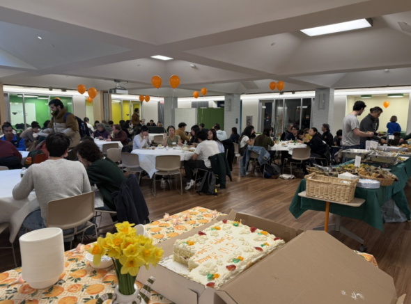 A huge meeting room with a round table covered by white tablecloths scattered around. Neon orange balloons are floating in the distance as people are immersed in the food and community!