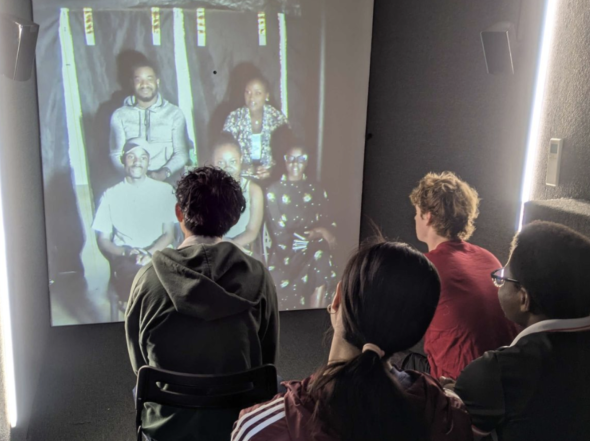 A group of Dartmouth students sit in an enclosed room with ultra-bright light on the wall. In front of them is a giant screen that's projecting live footage of students at Rwanda.