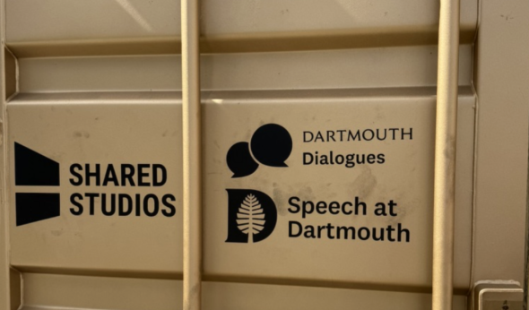 A metallic wall that has three printed logos: Shared Studios, Dartmouth Dialogues, and Speech at Dartmouth