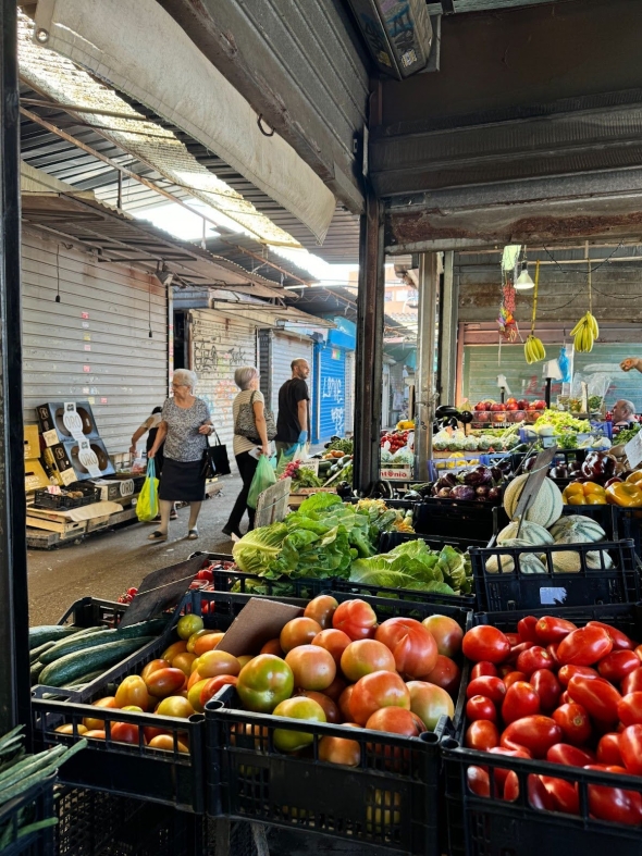 In the foreground, fresh produce sits in black bins at an open-air market. In the background, shoppers and vendors conduct their daily business.