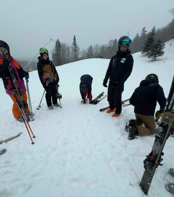 A group of people dressed in ski gear stands in a circle getting ready to put on their skis for skiing.