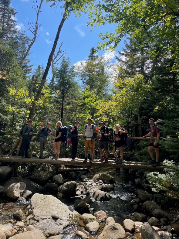  An image of me, Crystal, and my trip group outside on a wooden bridge. We are all carrying large frame packs on our backs. The background is a forest area and the bridge is over a small river.