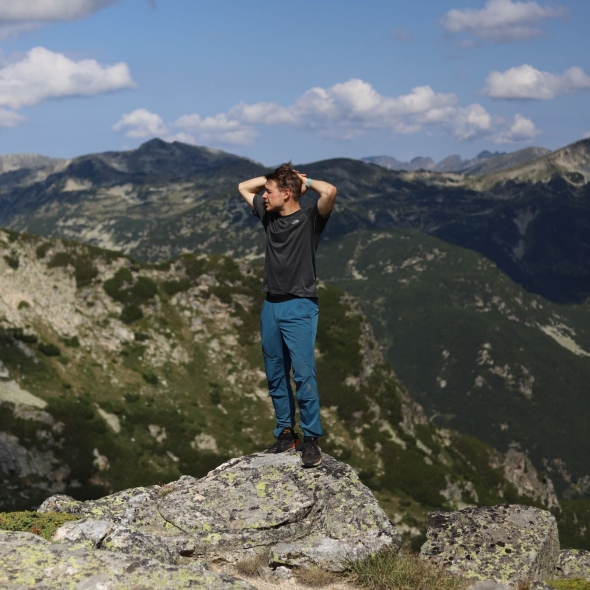 Me standing on a rock in dark blue pants and black T-Shirt with mountains in the background.