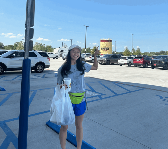 An image of me, Crystal, in a parking lot at Bucc-ee's, a gas station in Texas. I am motioning towards a large barrel of Bucc-ee's brand barbeque sauce in the background and carrying a plastic grocery bag of food.