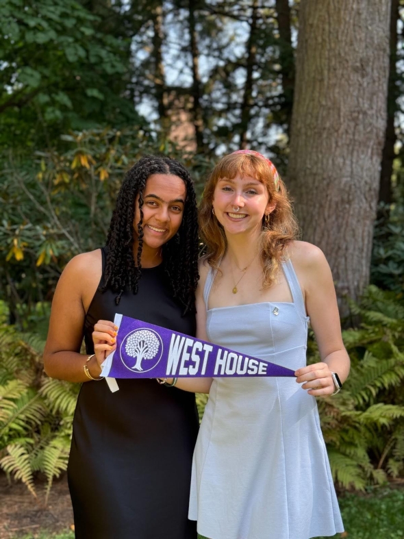 Two smiling girls—one in a white dress and one in a black dress—holding a purple pennant flag that says "West House" with the logo of a tree next to it. There are ferns and trees in the background.