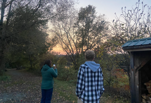 The sun is setting through the sparse treeline. Two teenagers, one wearing a green puffer jacket and the other wearing a black and white checkered sweatshirt, stand in the foreground of the image. There are trees on the left and the edge of a wooden shed