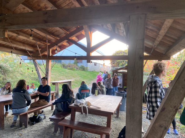 A group of twenty people gather under a wooden roof. Some sit on wooden picnic tables while others stand. The background is a green hill.