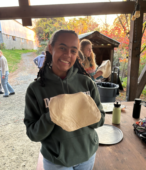 A girl wearing a green sweatshirt stands in the center of the frame. She is stretching out pizza dough over her knuckles. In the background, other people are doing similar things.