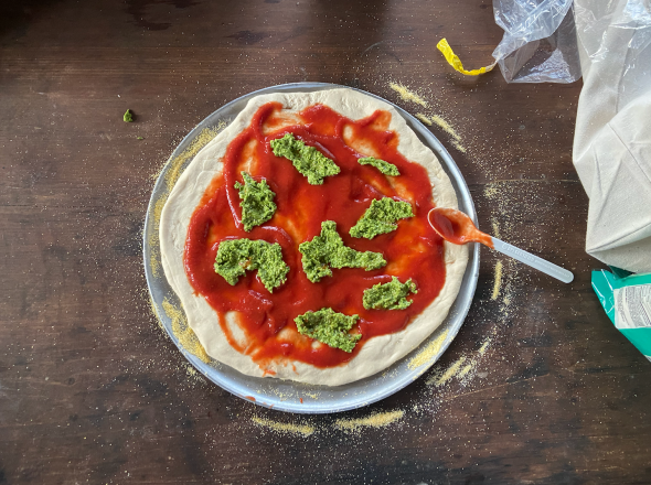 Pizza dough is rolled out on a pan. Red marinara is evenly spread on top of the dough. There are eight dollops of green pesto scattered onto the marinara. The white spoon that was used to spread the marinara still touches the pizza and is on the right