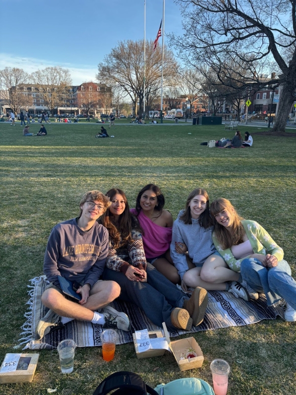 4 girls and 1 boy outside on the green sitting on a picnic blanket smiling.
