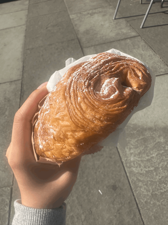 An image of me, Crystal, holding a croissant. The croissant is covered in powdered sugar and the background is of the concrete ground.