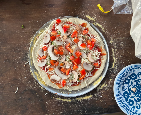 An uncooked pizza is on a silver pizza pan. The pizza has red marinara sauce, mushrooms, red bell peppers, and mozzarella cheese on top of it. On the lower right-hand side, there is a small plate with a blue pattern on top.