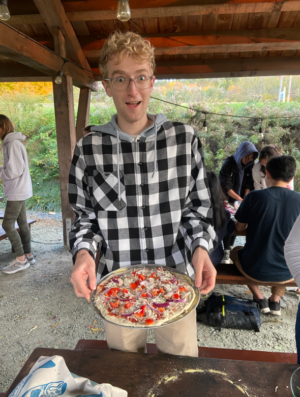 A tall man with blond hair is wearing a black and white checkered sweatshirt. He is standing in the middle of the photo, holding an uncooked pizza. The pizza has bell peppers, mushrooms, onions, cheese, and sauce on it.