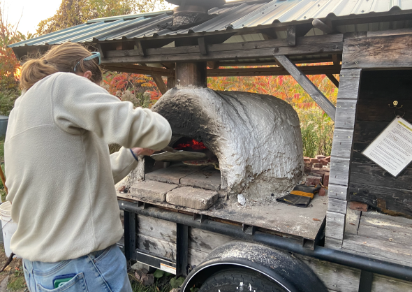 A girl in a cream-colored sweatshirt stands in front of a small pizza oven. The oven is on a wooden cart with a metal roof. The oven itself appears to be made out of clay.