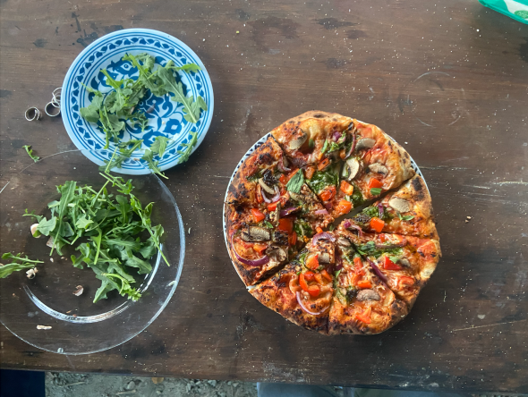 A cooked pizza is cut into eight slices. On the left of the pizza, there are two plates holding some arugula. One plate is white with a blue pattern, and the other is clear. The plates and the pizza are all sitting on a wooden table with a dark stain.