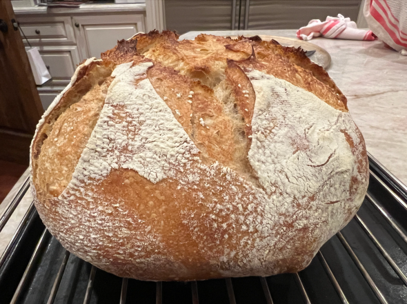 A round loaf of sourdough bread with scores on the top takes up the majority of the image. The bread is dark tan/light brown with flour dusted on top. The loaf sits atop a silver wire rack and a dark gray tray.