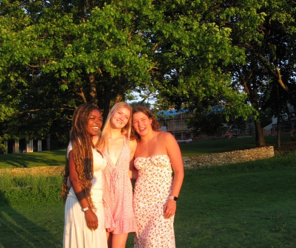 Three friends smiling at the camera, wearing dresses, in the midst of a grassy field. 