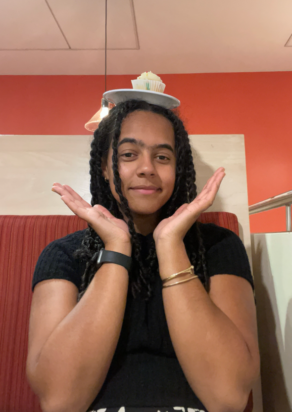 A girl wearing a black shirt balancing a small white plate on her head. On top of the plate is a cupcake. The girl is holding her hands up to form a V under her chin.