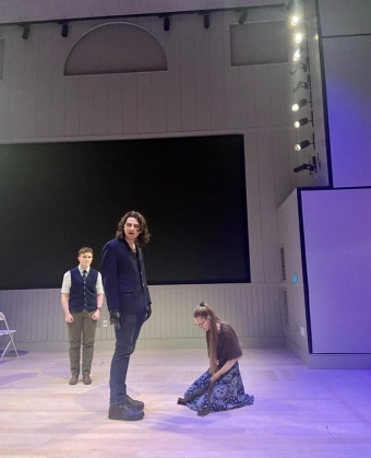 three actors on a stage lit in purple lighting. A woman on her knees, mournful. a man in black, scowling but contemplative. a men in the background, hesitant and waiting for the first man's reaction.