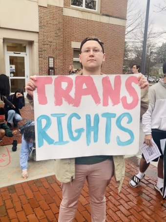 A student is standing, looking at the camera, holding a poster that says "trans rights." "trans" is written in light pink, "rights" is written in light blue.