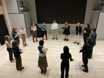 Richard II Pre-Show Warm-Up Circle A group a students stand in a circle, evenly spaced. They are wearing costumes, primarily modern formal wear.