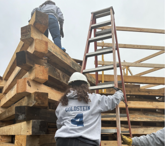  Serena climbs up a yellow adder in the center of the picture. She wears a gray sweatshirt and around her is the yellow wooden beams of the bonfire. 