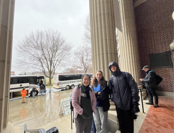 Three students stand near each other with smiles. Tall columns are on either side of them.