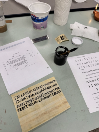 A papyrus with Latin written on it in black ink sitting on a table. There is a jar of ink and various other papers on the table as well.