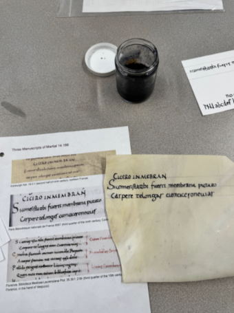 Parchment with Latin written on it in black ink sitting on a table. There is a jar of ink and various other papers on the table as well.
