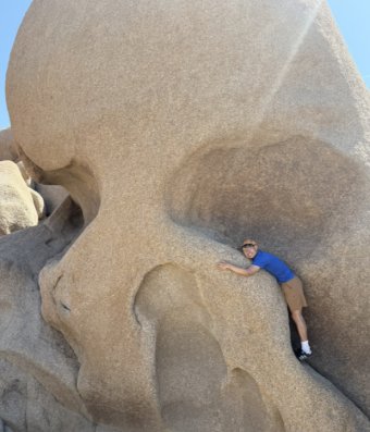 A man in shorts and blue shirt is hugging a huge boulder shaped like a skull.